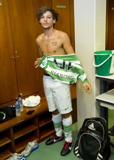 a shirtless man standing in a dressing room holding a bag and soccer boots on the floor