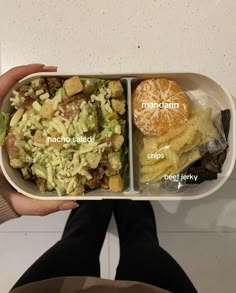 a person holding a container with food in it that includes salad, fruit and an orange