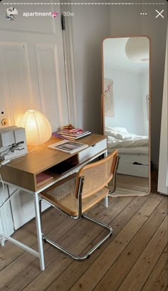 a desk with a mirror, lamp and bed in the background on a wooden floor