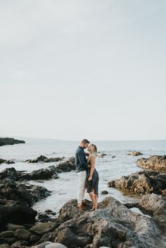 a man and woman standing on rocks near the ocean with their arms around each other