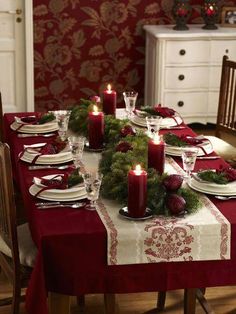 the table is set for christmas dinner with candles on it and red napkins, surrounded by greenery