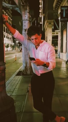 a man in white shirt and black pants standing next to a street pole holding a cell phone