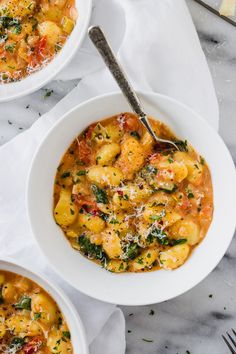 two white bowls filled with pasta and vegetables on top of a marble table next to silverware