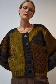 a woman standing in front of a white wall wearing a green and brown sweater with buttons