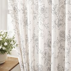 a vase with white flowers sitting next to a curtain in front of a window on top of a wooden table