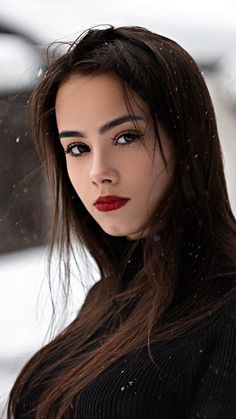 a woman with long brown hair and red lipstick is standing in the snow wearing a black sweater