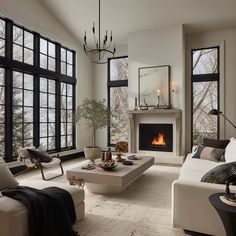 a living room filled with white furniture and large windows next to a fire burning in the fireplace