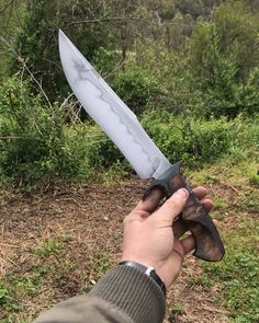 a hand holding a knife in the middle of a field with trees and bushes behind it