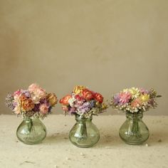 three green vases filled with colorful flowers sitting on a white table top next to each other