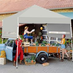 three people standing under a blue tent next to a truck with vegetables on the back