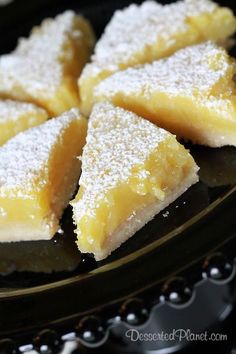 several pieces of lemon bars are on a black plate with powdered sugar over them