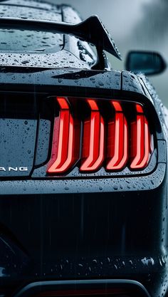 the rear end of a black sports car with red tail lights and rain drops on it