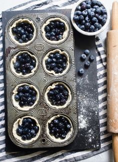 blueberry pies in a muffin tin next to a rolling pin and bowl of blueberries