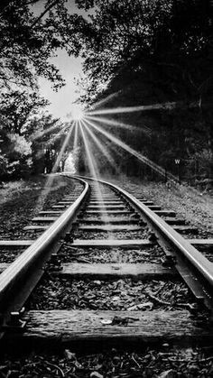 black and white photograph of train tracks with the sun shining through trees in the background