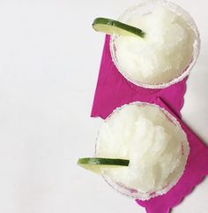 two margaritas with cucumber and sugar in them on a pink napkin next to each other