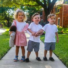 three little kids standing on the sidewalk holding hands with each other and smiling at the camera