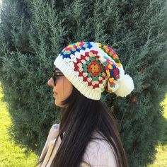 a woman standing in front of a bush wearing a crocheted hat with pom poms