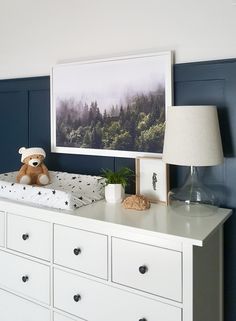 a teddy bear sitting on top of a white dresser next to a painting and lamp