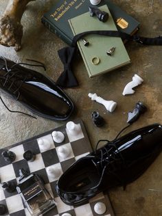 An artistic arrangement of groom accessories including glossy black dress shoes, a black bow tie, cufflinks, a wedding band, and a luxury cologne, all placed on a chessboard with vintage books, symbolizing elegance and sophistication for a luxury wedding. Wedding Accessory Photos, Groom Detail Shots, Groom Idea, Sailboat Elopement, Creative Flatlay, Nyc Editorial, Weddings 2024