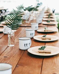 a long wooden table with white dishes and plates on it, along with greenery