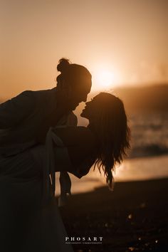 a man and woman kissing on the beach at sunset with the sun in the background