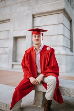 a man in a red graduation cap and gown sitting on a stone bench with his legs crossed