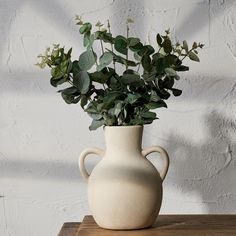 a white vase filled with green leaves on top of a wooden table