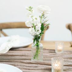 white flowers in a glass vase on a table with plates and candles around it,