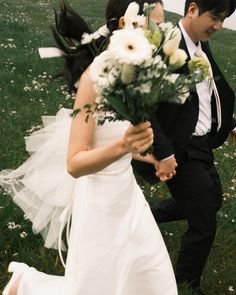 a bride and groom walking in the grass with flowers on their wedding day, holding each other's hands