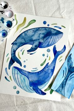 two watercolor paintings of blue whale's on paper next to paintbrushes