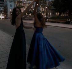 two women in long dresses are dancing on the sidewalk at night with their hands up