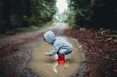 a little boy in red boots playing in the mud on a dirt road surrounded by trees