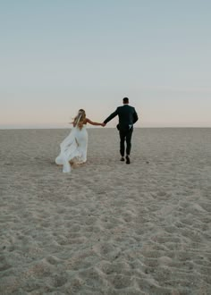 a bride and groom holding hands while walking in the sand at sunset on their wedding day