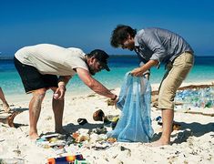 two men picking up trash on the beach, one is holding a blue mesh bag