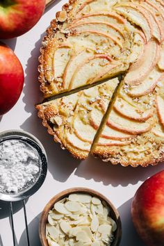 an apple pie with slices cut out next to some apples and other ingredients on a white surface