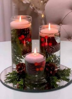 three lit candles sitting on top of a glass plate covered in greenery and pine cones