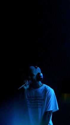 a man standing in the dark holding a microphone up to his ear and wearing a white t - shirt with words on it