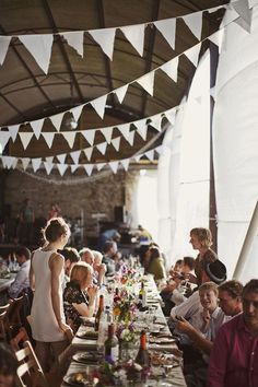 people are sitting at a long table with food on it and bunting above them