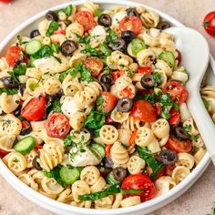 a white bowl filled with pasta salad next to some tomatoes and cucumber slices