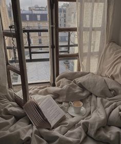 an open book and cup sit on a bed in front of a window overlooking the city