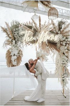 a bride and groom kissing under an arch decorated with white flowers, greenery and feathers