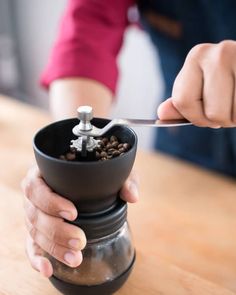 a person holding a spoon over a coffee grinder on top of a wooden table