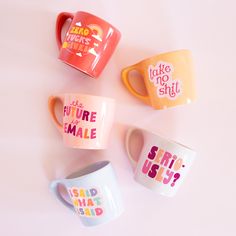 four coffee mugs with different sayings are on a pink surface and one is white