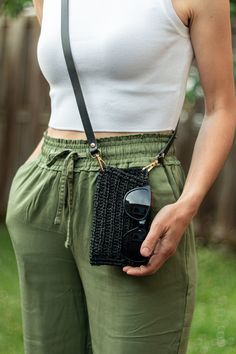 a woman in green pants holding a cell phone and wearing a white tank top with black straps