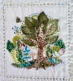 an embroidered tree with leaves and flowers on a white quilted tablecloth, next to a bee