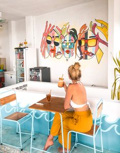 a woman sitting at a table with a drink in her hand and paintings on the wall behind her