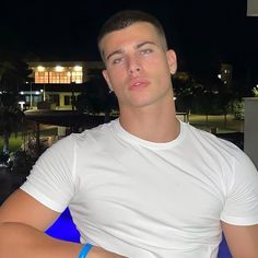 a man in white shirt and blue bracelets sitting on a chair with his arms crossed