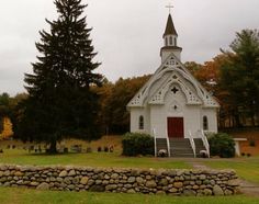 a white church with a red door and steeple surrounded by trees on a cloudy day