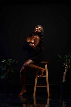 a woman sitting on top of a wooden stool in front of a black wall and potted plant