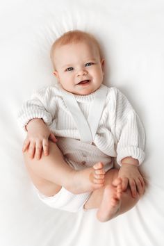 a baby is smiling while sitting on the bed with his legs spread out and feet crossed